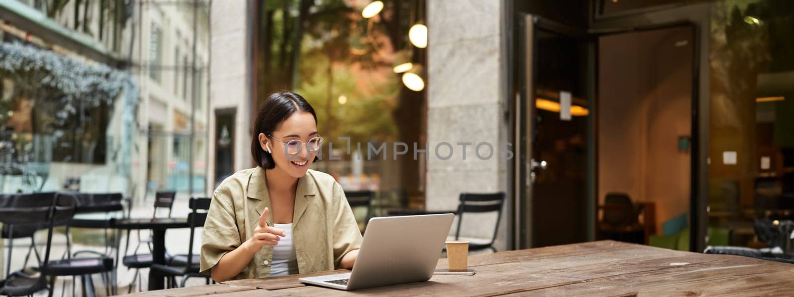 Young woman sitting on online meeting in outdoor cafe, talking to laptop camera, explaining something, drinking coffee.