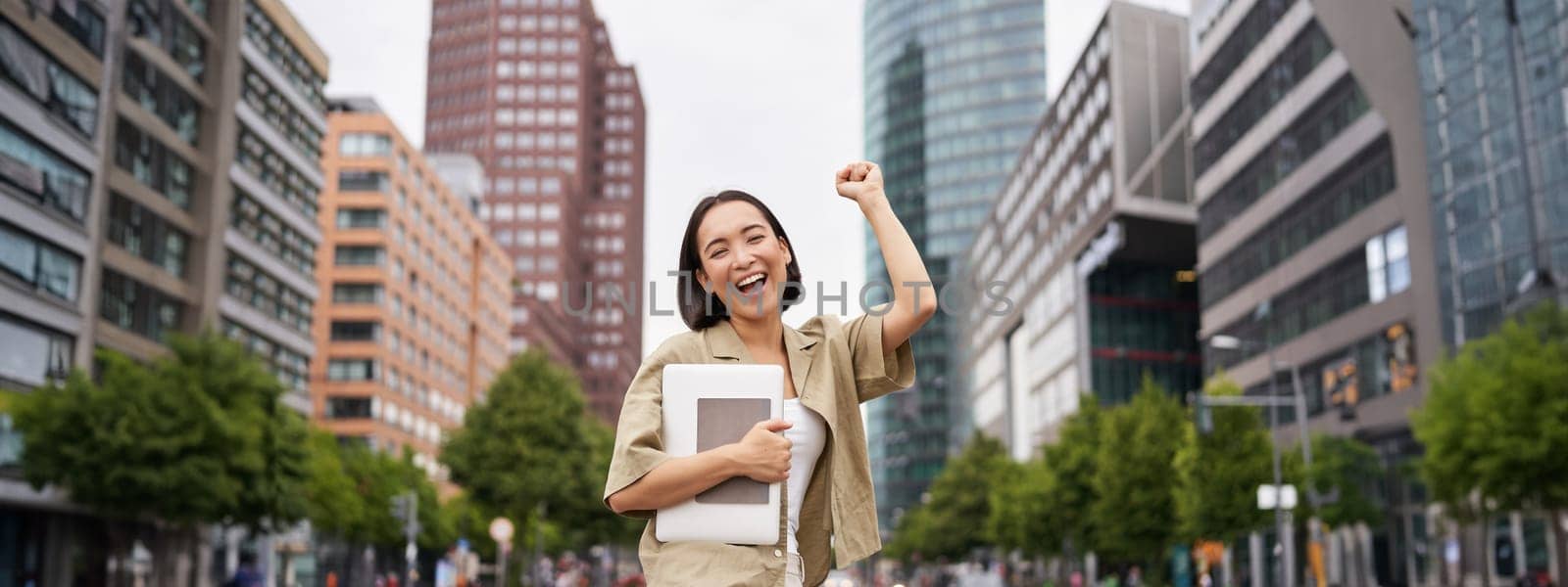 Portrait of happy asian woman stands with tablet near street road, cheering, raising hand up in triumph, celebrating by Benzoix