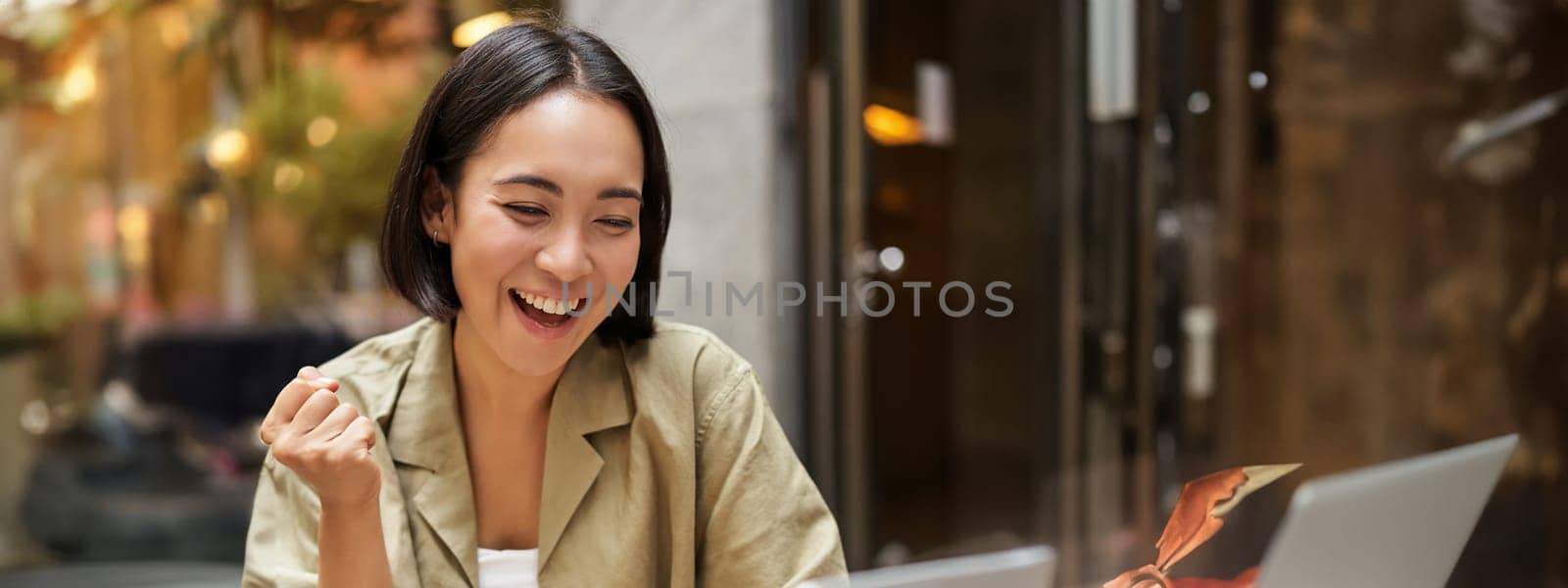 Vertical shot of happy girl talking on video call, looks at laptop, having online meeting, sitting in outdoor cafe.