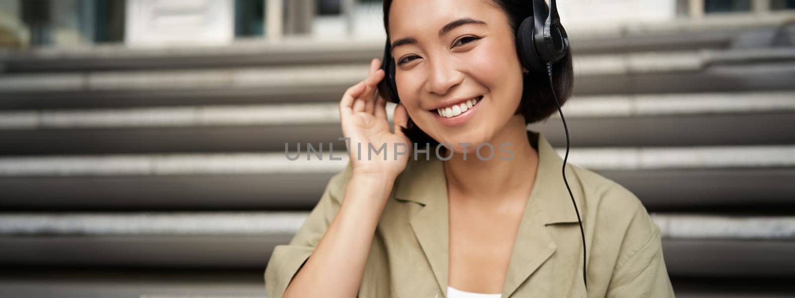 Happy asian girl listens to music in headphones, smiles, sits outdoors on stairs by Benzoix