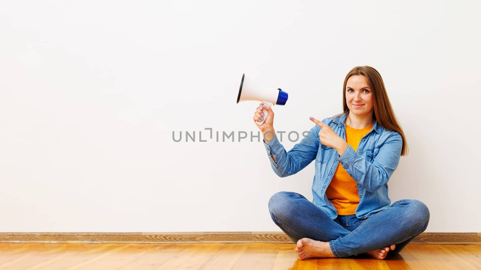 Young beautiful woman with megaphone sitting on wooden floor in front of white wall, copy space.
