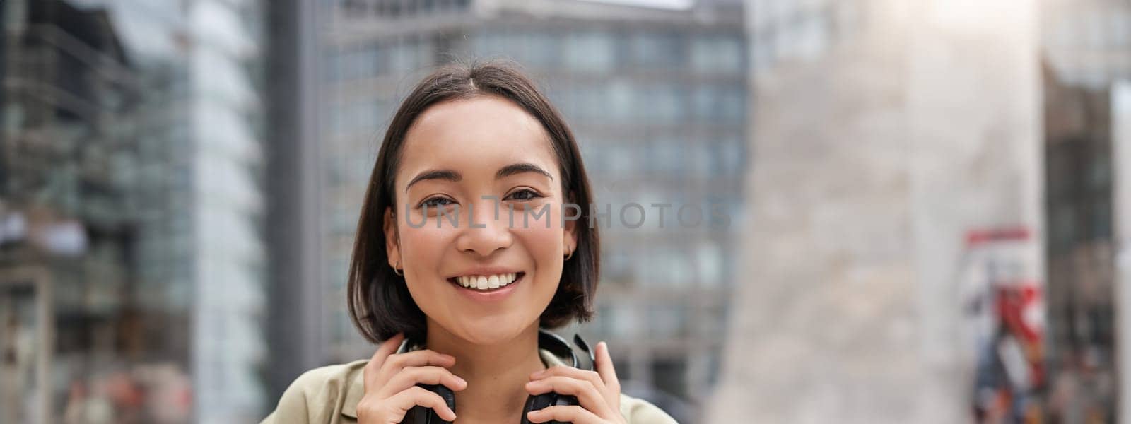 Vertical shot of beautiful asian woman posing with headphones around neck, smiling and laughing, standing on street in daylight.