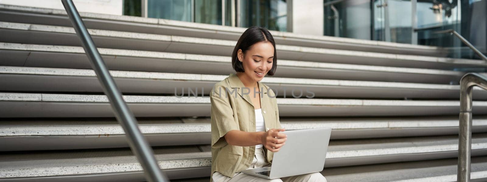 Portrait of young woman student, asian girl using laptop. Asign smiling girl, digital nomad works on her project remotely, sitting on street.