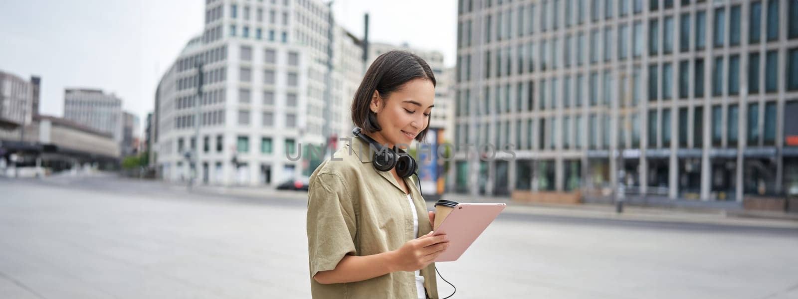 Asian girl walking on street, reading on tablet and drinking coffee outdoors.