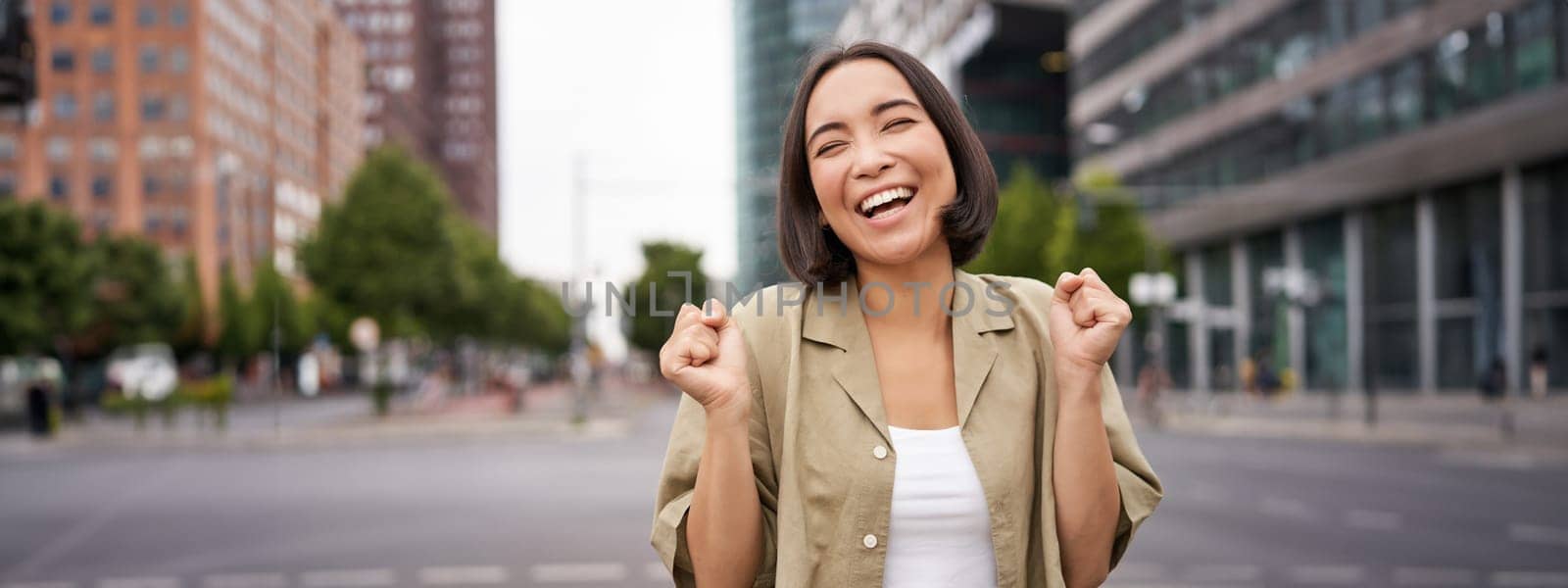 Happy asian girl triumphing on streets of city, dancing from happiness, celebrating victory, posing outdoors.