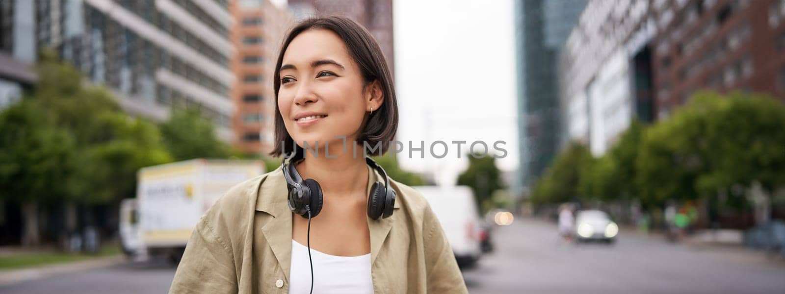 Portrait of young asian woman in headphones, posing in city, smiling and looking away, standing on street of city centre.