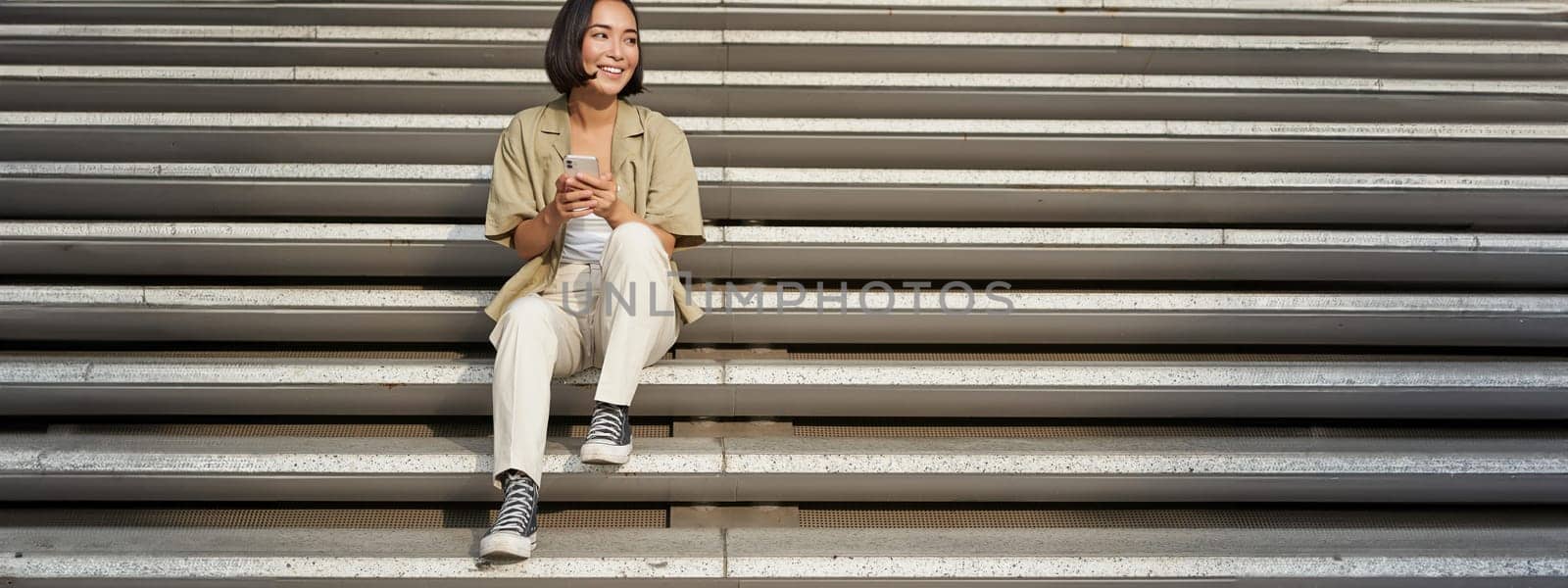 Portrait of smiling asian girl sits on stairs with her smartphone, browsing internet on mobile phone, resting outdoors in city centre by Benzoix