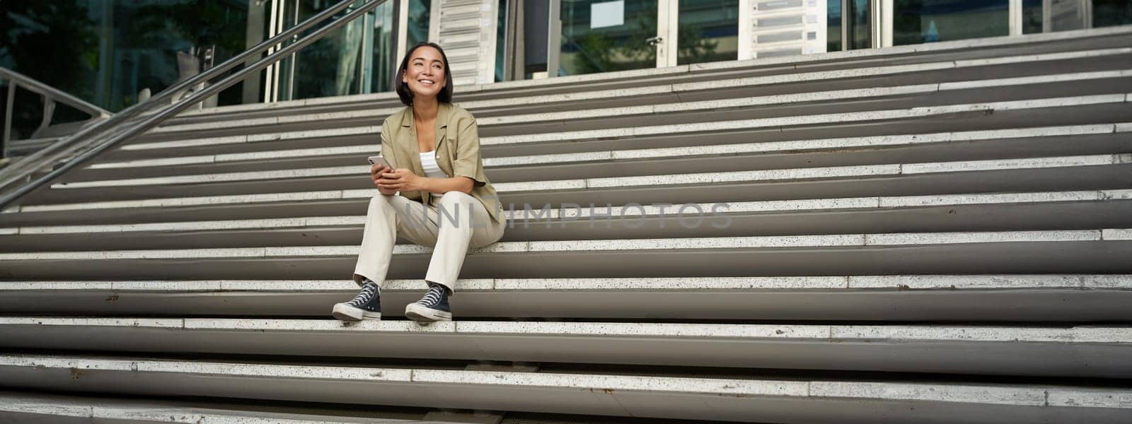 Portrait of smiling asian girl sits on stairs outdoors, sending message, using smartphone app, looking at mobile phone screen by Benzoix