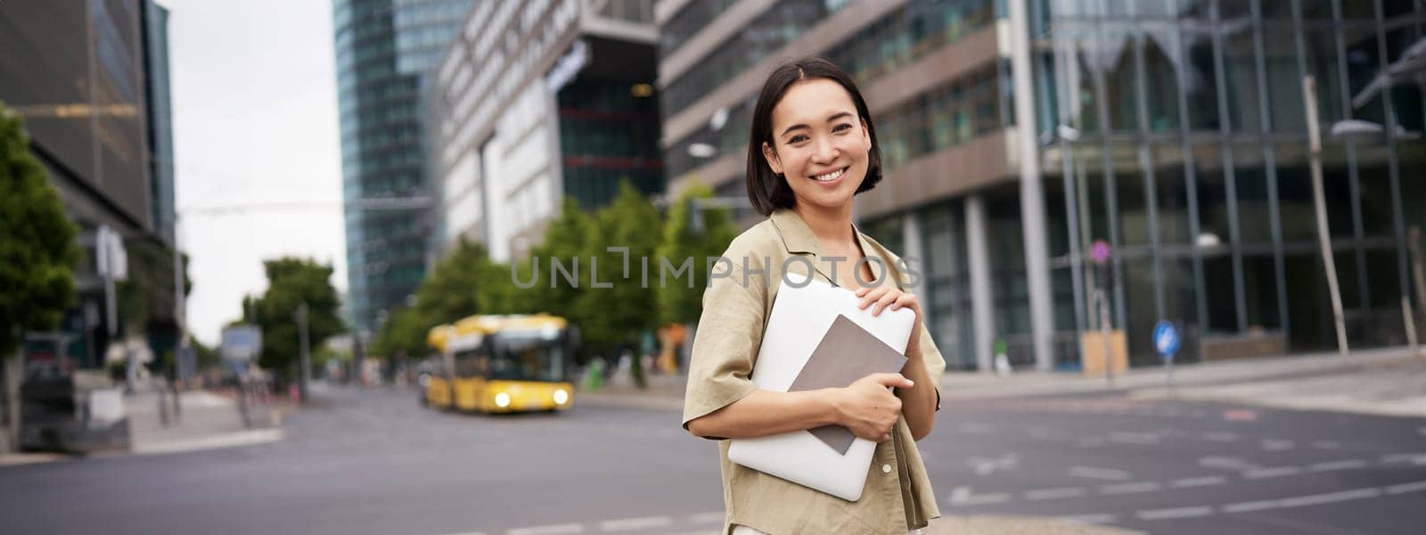 Beautiful asian girl smiles as commutes to work, stands on street with laptop and notebook.