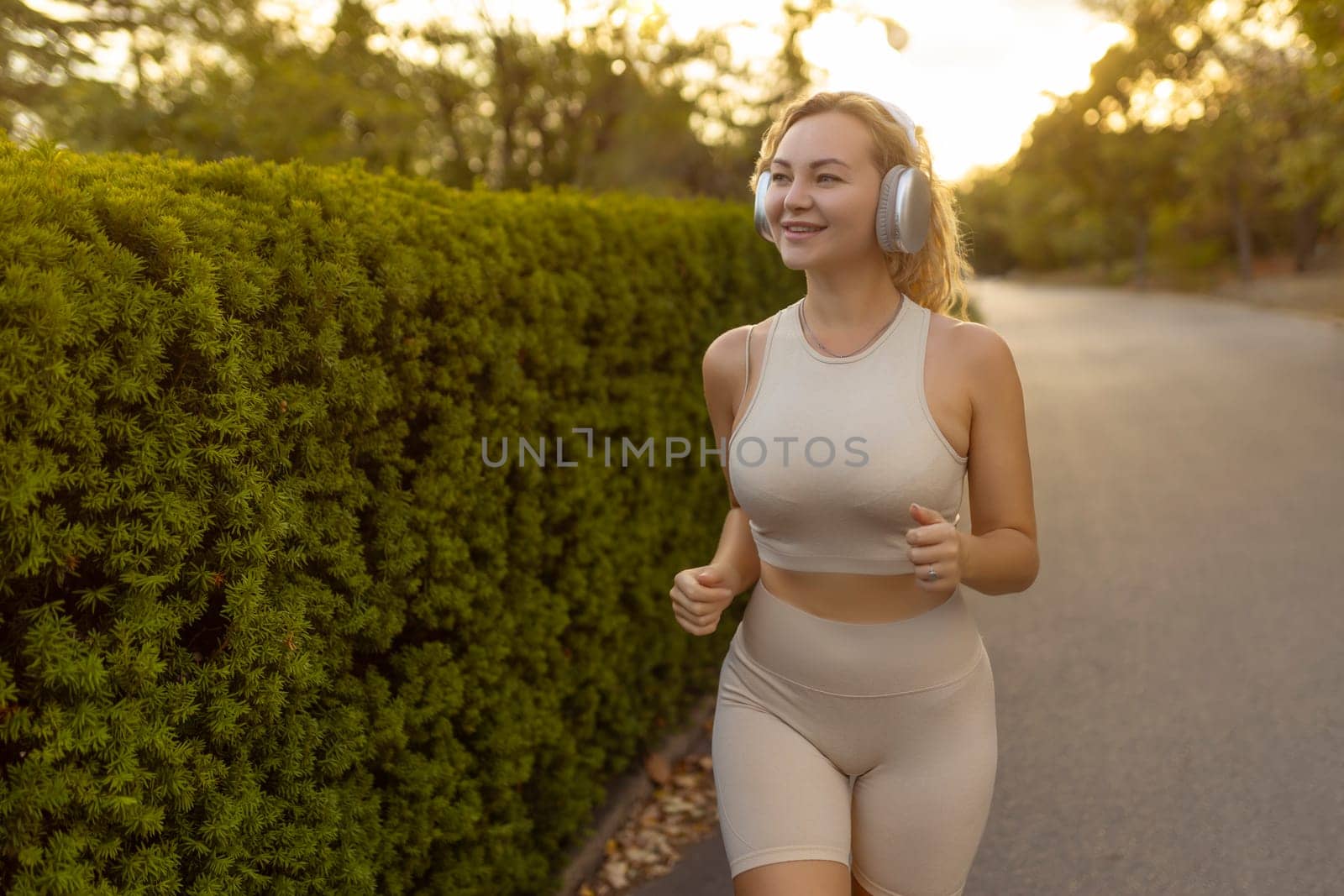 Smiling blonde woman wearing modern wireless headphones running through the park on a bright sunny summer day. A happy girl with a sweet smile runs and listens to good music. Lifestyle.