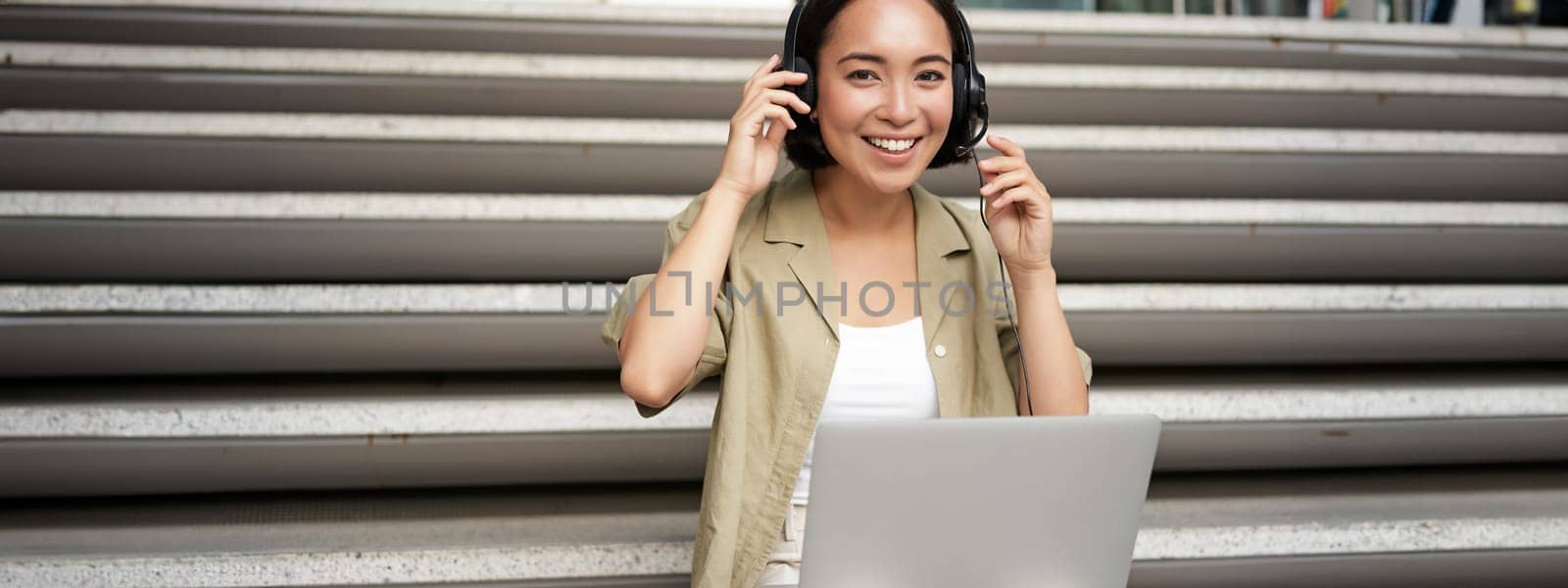 Smiling asian girl in headphones, works on laptop, digital nomad using computer on remote, sitting on stairs outdoors by Benzoix