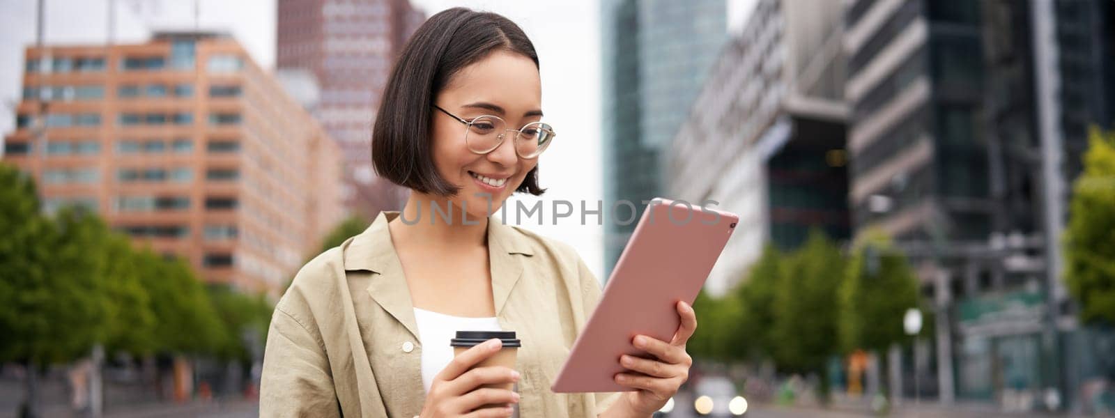 Woman standing on street, drinking coffee, reading on tablet, checking messages, using app.