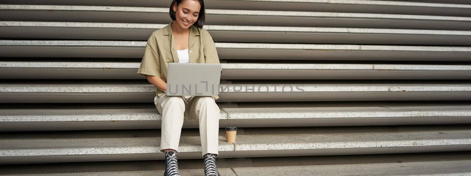 Portrait of young asian woman using her laptop, sitting outdoors on stairs. Happy smiling girl with personal computer.