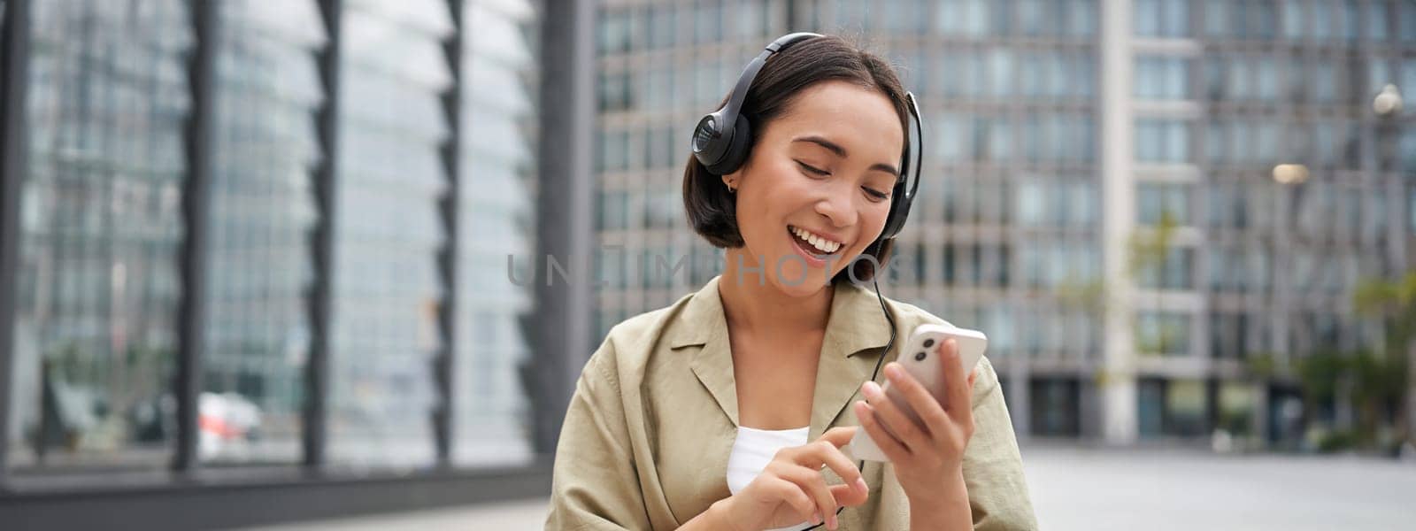 Happy girl dances on street and listens music in wireless headphones, holds mobile phone, using streaming app.