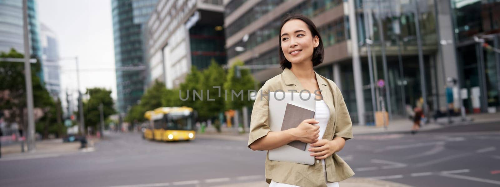 Beautiful asian girl smiles as commutes to work, stands on street with laptop and notebook.