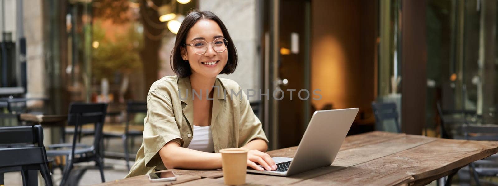 Portrait of smiling girl in glasses, sitting with laptop in outdoor cafe, drinking coffee and working remotely, studying online by Benzoix