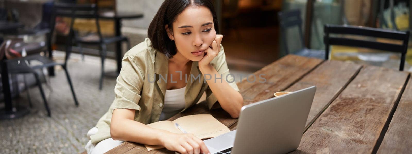 Bored asian girl sitting in cafe and staring at laptop, studying and feeling sad, doing homework by Benzoix