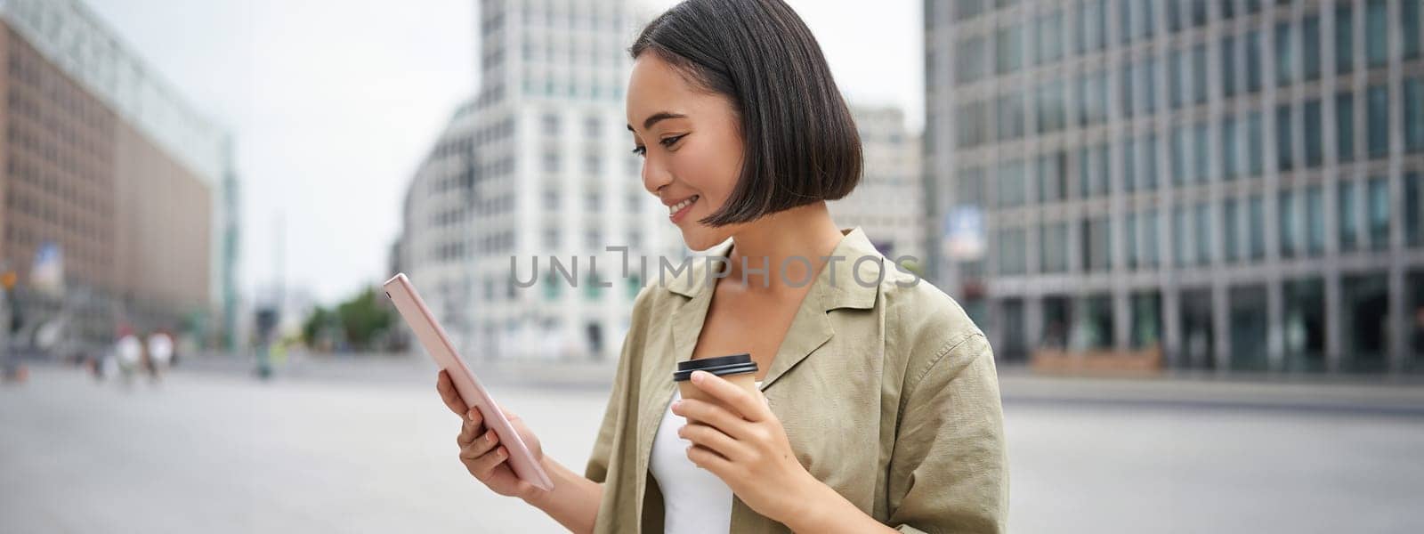 Portrait of smiling korean girl looks at tablet, reads on street, drinks takeaway coffee.