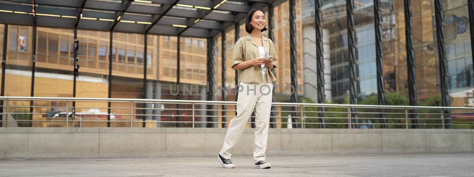 Portrait of young asian woman, student with smartphone, standing on street near glass building, waiting for someone by Benzoix