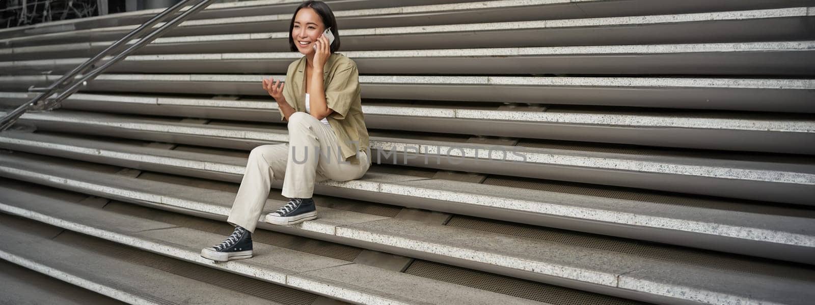 Smiling asian girl sits on stairs of building and talks on mobile phone, relaxing during telephone conversation.
