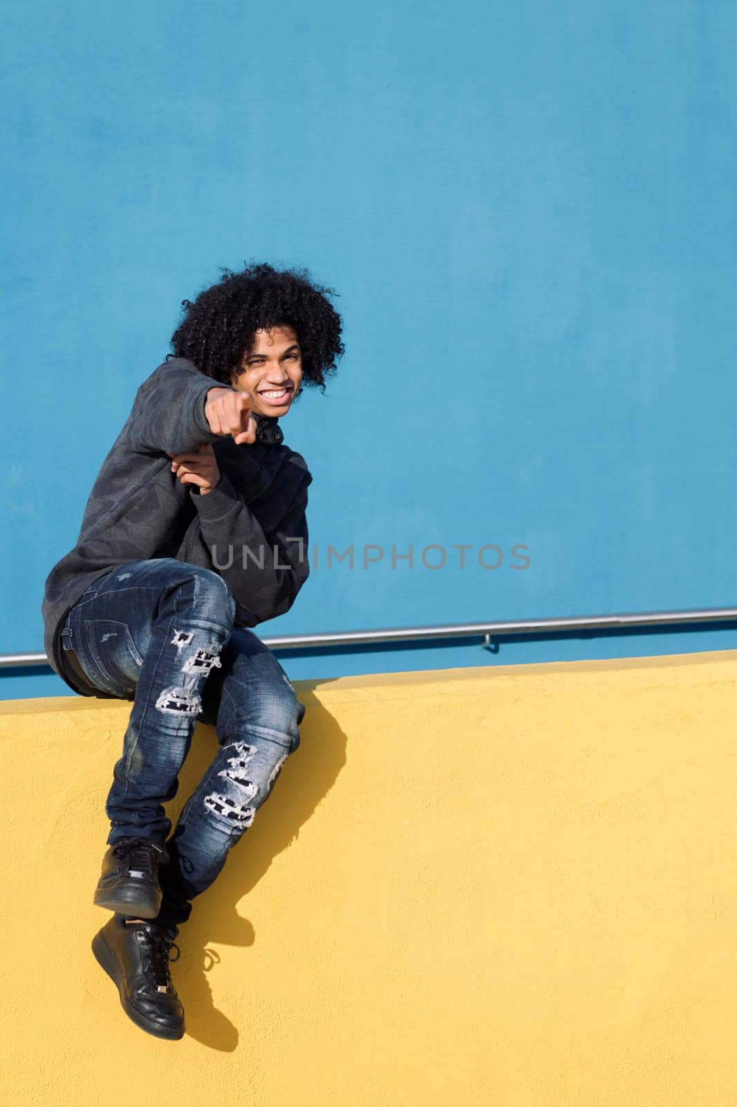 funny african american teenager smiling happy pointing looking at camera while sitting on a yellow wall in front of blue background, concept of adolescence and young lifestyle, copy space for text