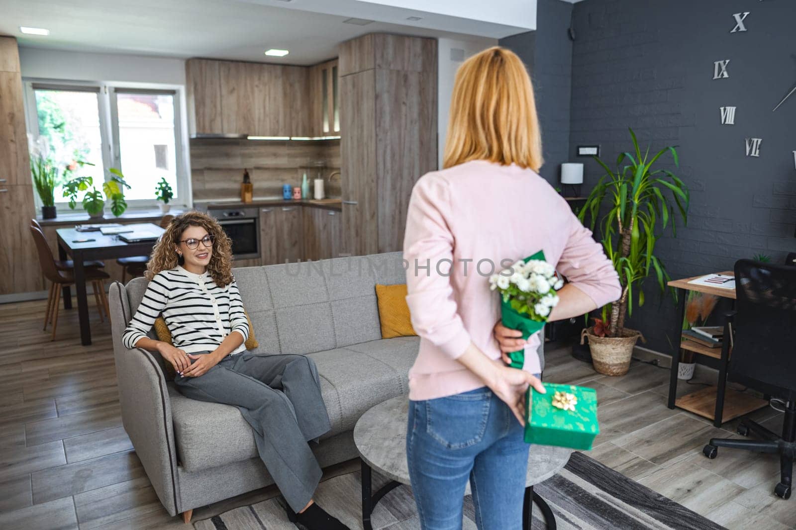 A blonde smiling woman surprises her best friend with a gift and flowers, standing in the middle of the living room hiding the gift and a white bouquet behind her back.