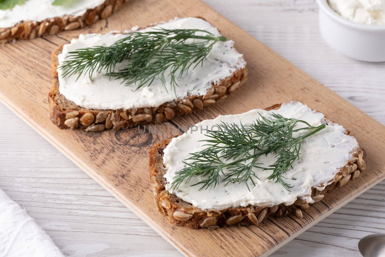 Two slices of rustic rye bread are generously spread with creamy topping, garnished with fresh dill, placed on a wooden board in a well-lit kitchen.