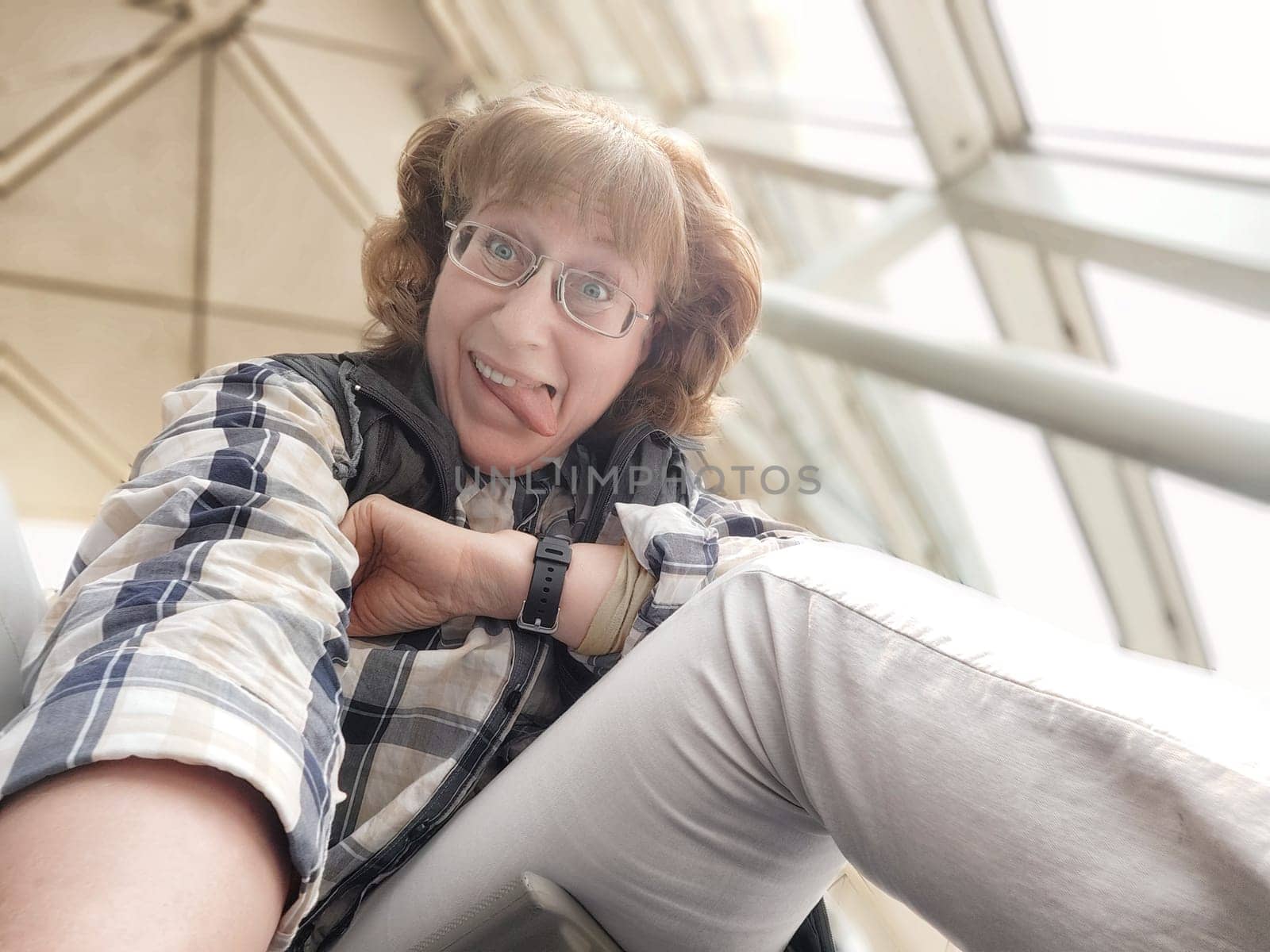 A woman in her middle ages takes a cheerful selfie in a busy airport hall while waiting for her flight. Her playful expression reflects her excitement for upcoming travels.