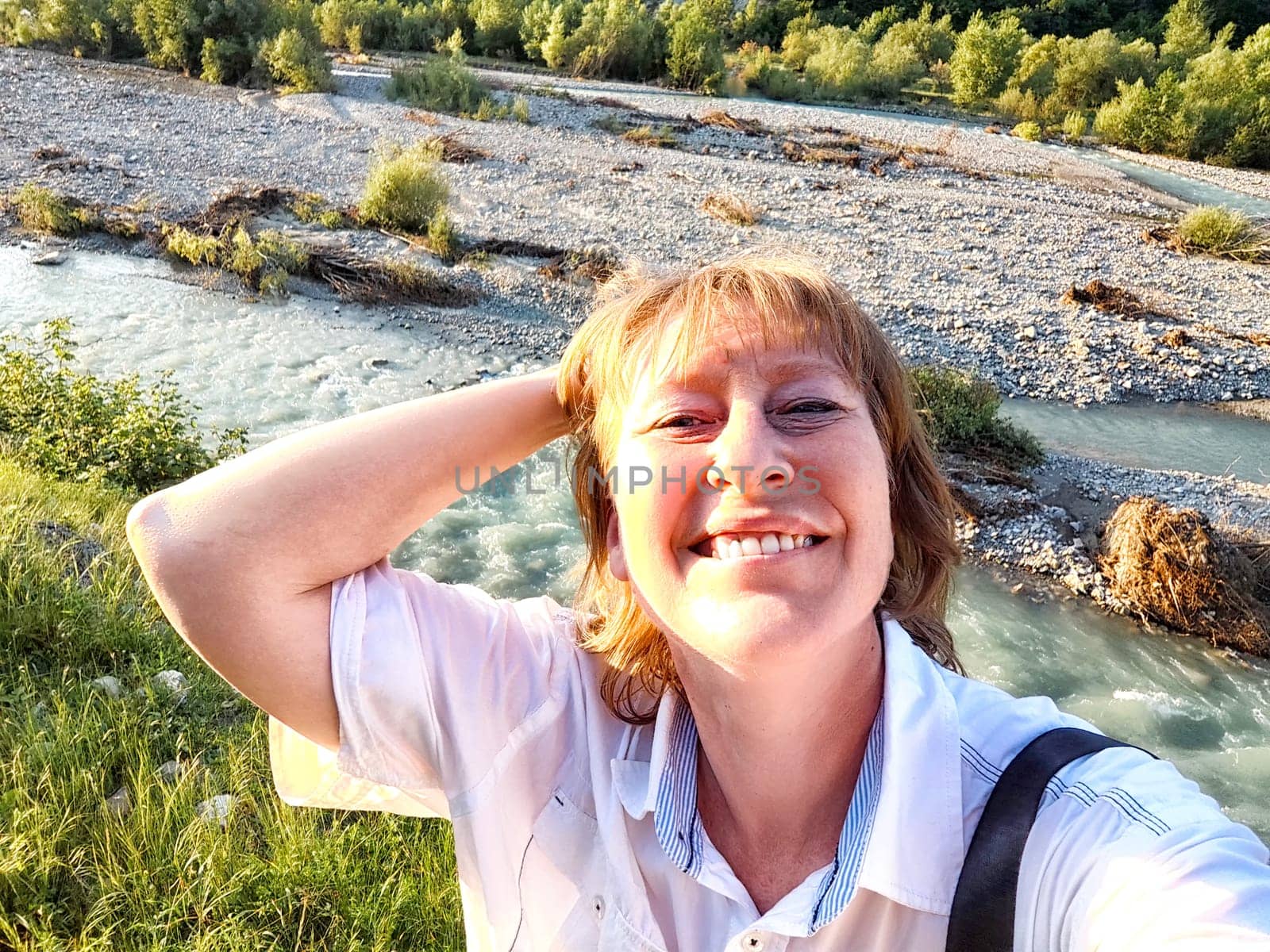 A tourist blogger joyfully captures a selfie near a rushing mountain river, surrounded by nature and rugged terrain