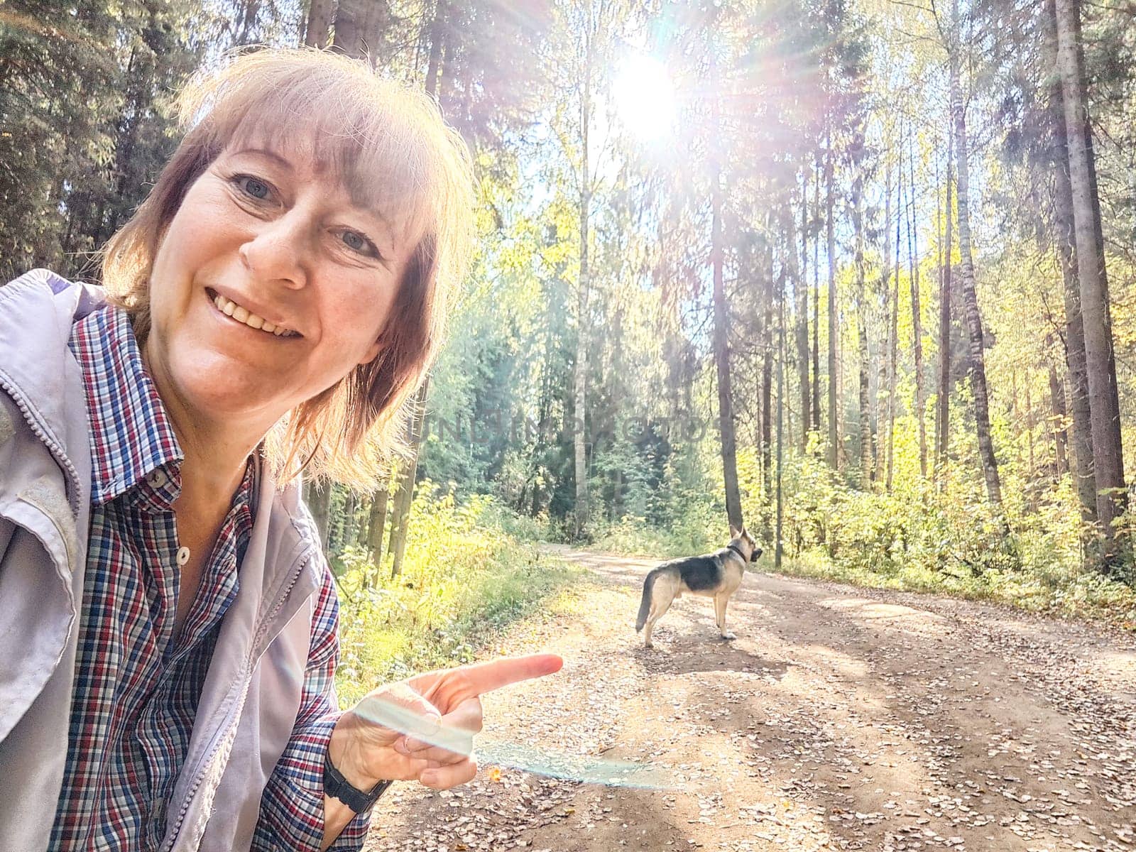 A middle-aged woman takes a joyful selfie while walking her dog along a sunlit dirt path in a lush forest.