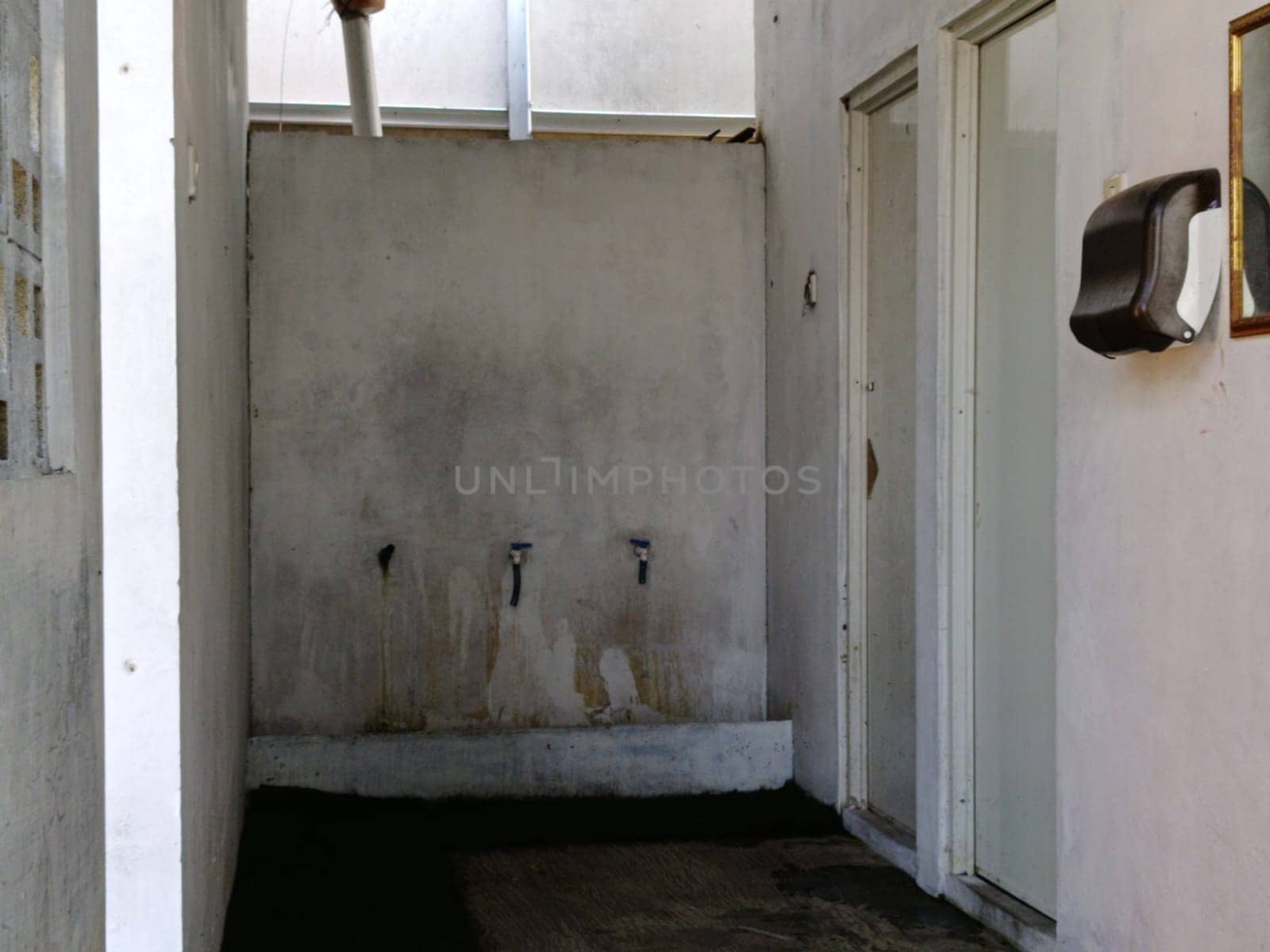 An old, neglected washroom with stained concrete walls and worn faucets. The image conveys a sense of abandonment and decay, portraying a stark, utilitarian environment.