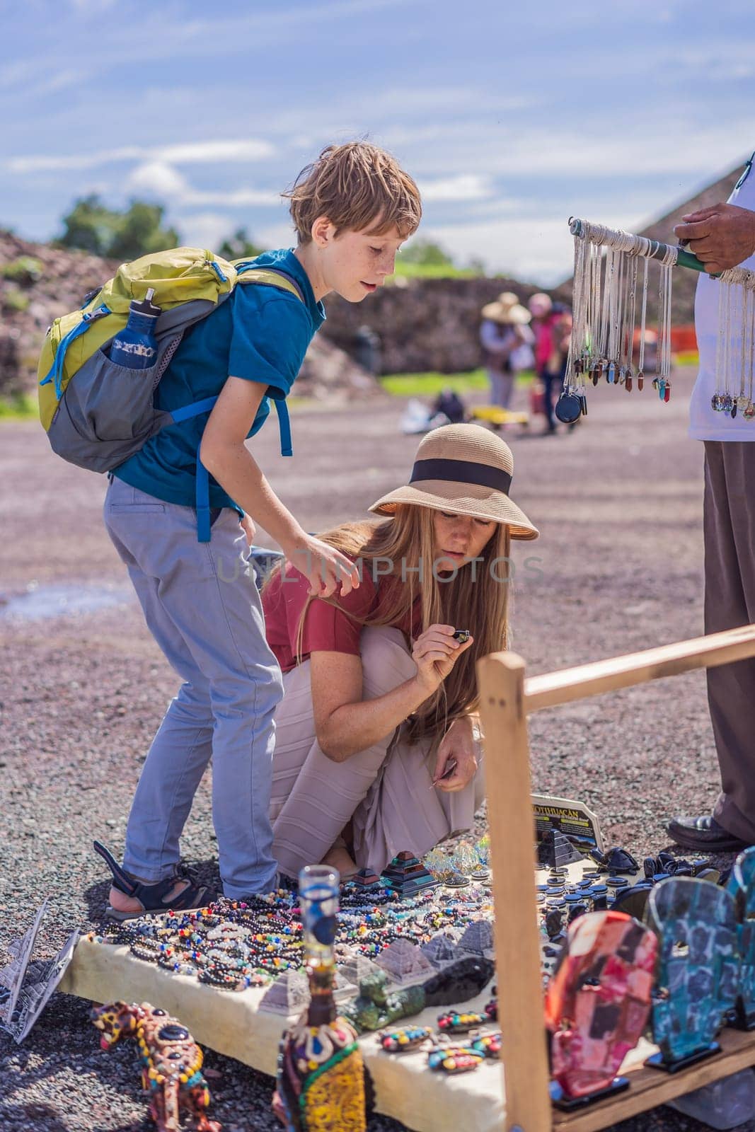 Mother and son tourists buying Mexican souvenirs from a street vendor at the foot of the Teotihuacan pyramid. Cultural experience, travel, and local craftsmanship concept.