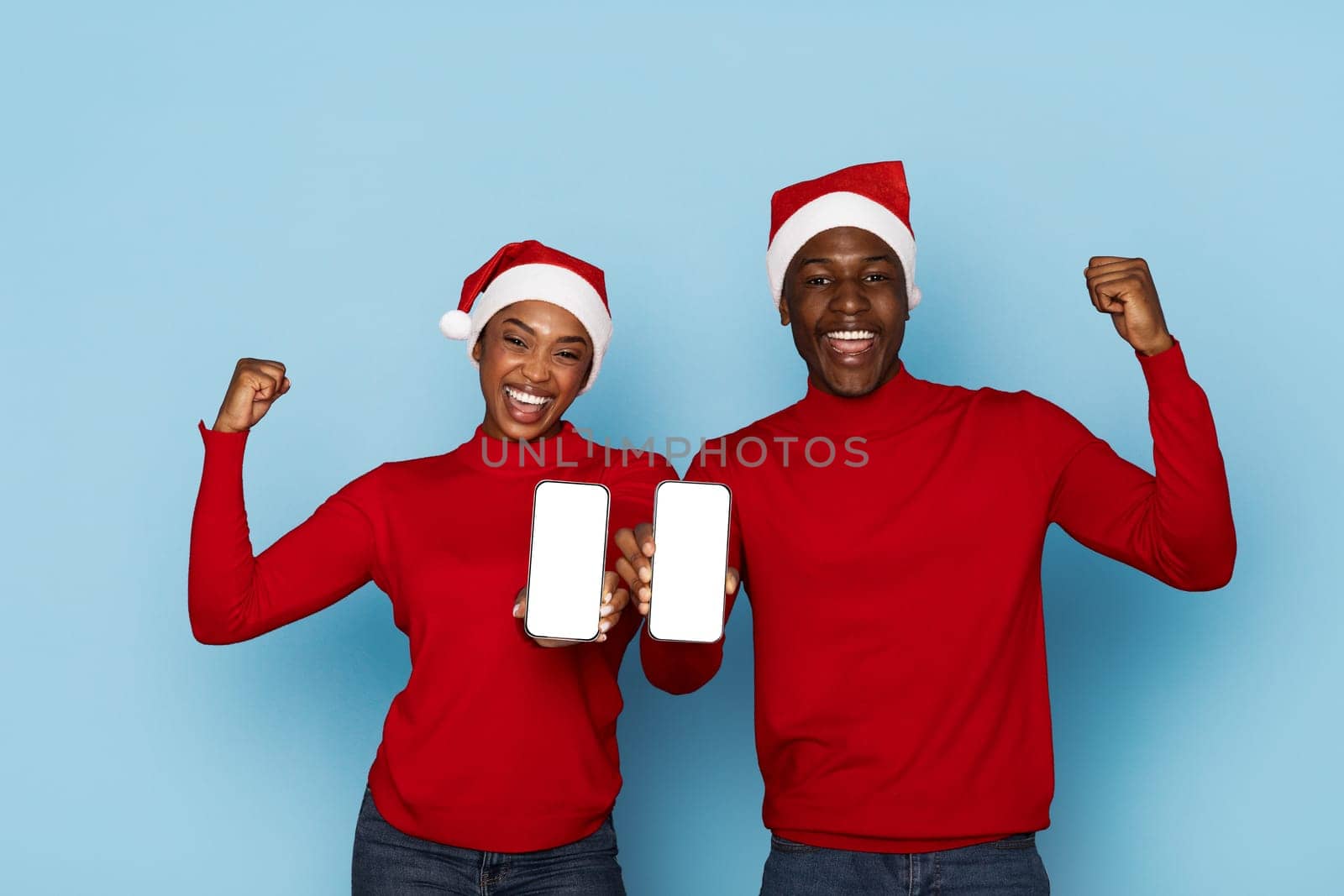 A joyful black couple, wearing matching red sweaters and Santa hats, proudly holds up their smartphones while smiling enthusiastically against a light blue background.