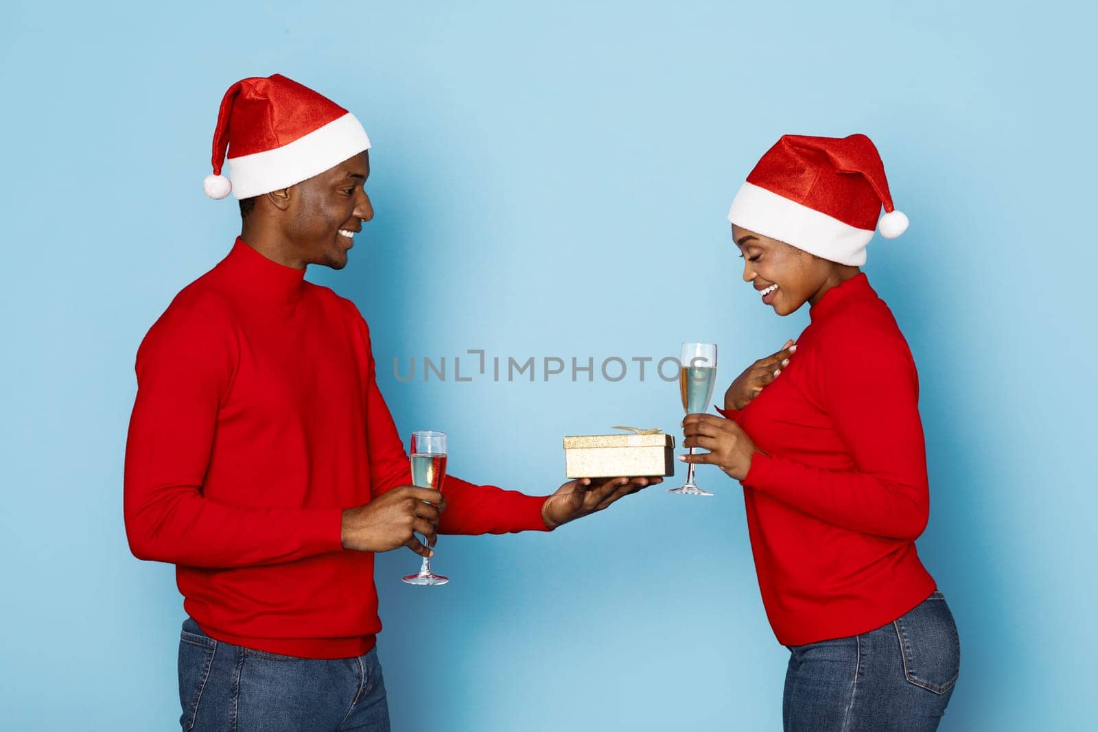 A joyful couple, both wearing festive red sweaters and Santa hats, exchanges a beautifully wrapped gift while holding glasses of champagne, capturing the spirit of Christmas together.