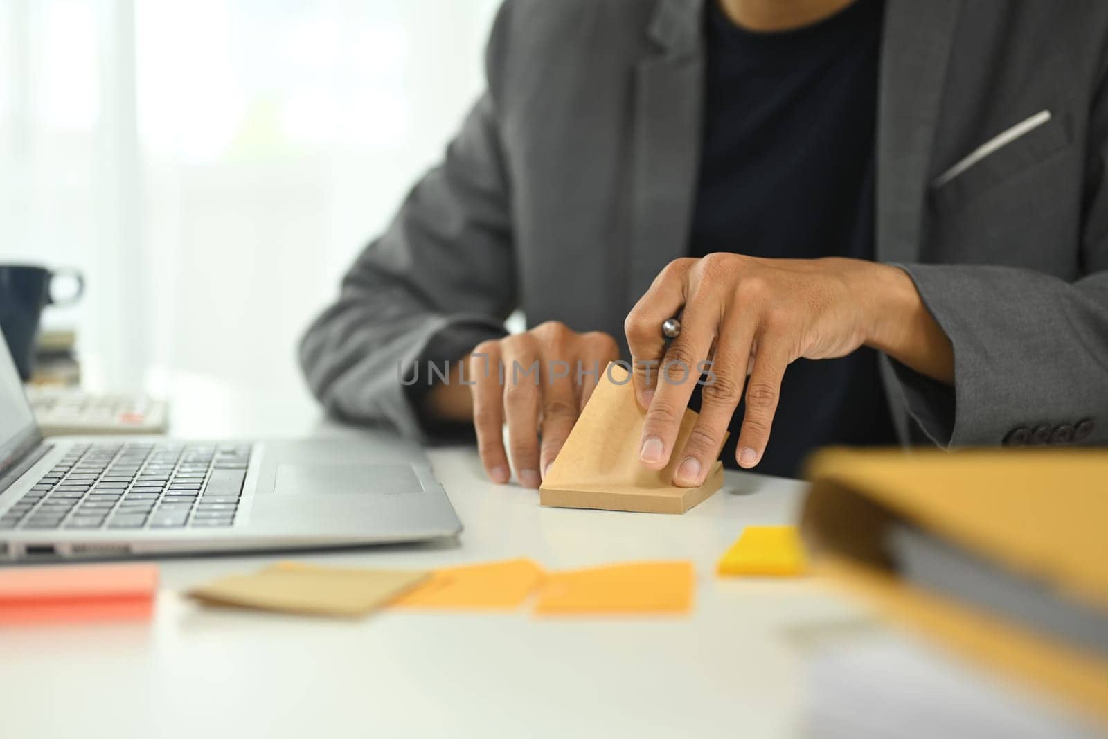 Unrecognizable sitting at a desk with a laptop and writing idea on adhesive note.