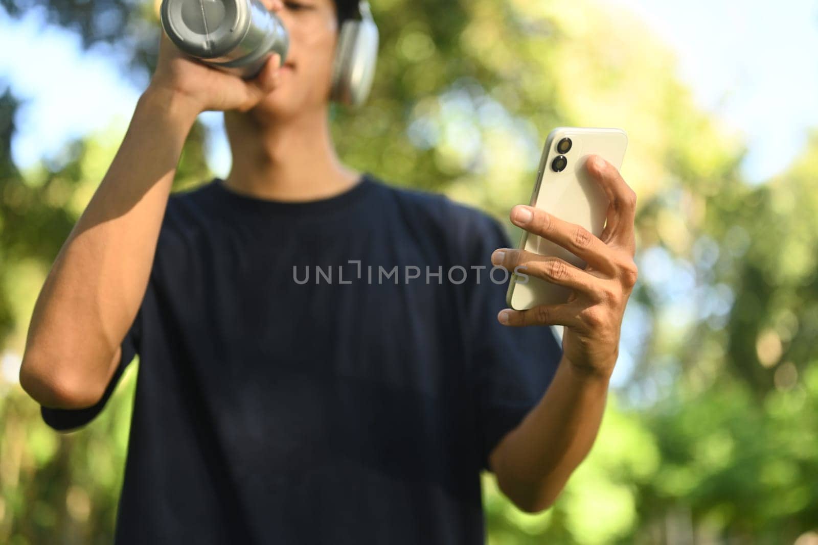 Young athletic man taking a break outdoors, drinking water and using mobile phone.
