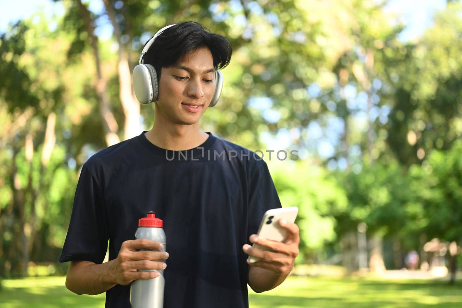 Young athletic man taking a break outdoors, drinking water and using mobile phone.