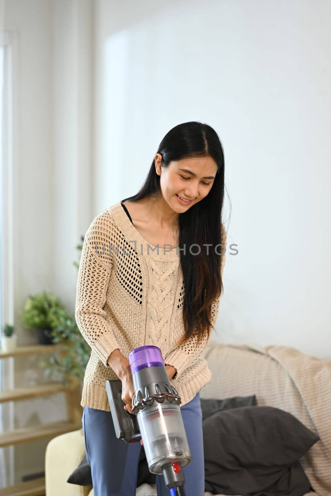 Young woman vacuuming couch surface with cordless vacuum.
