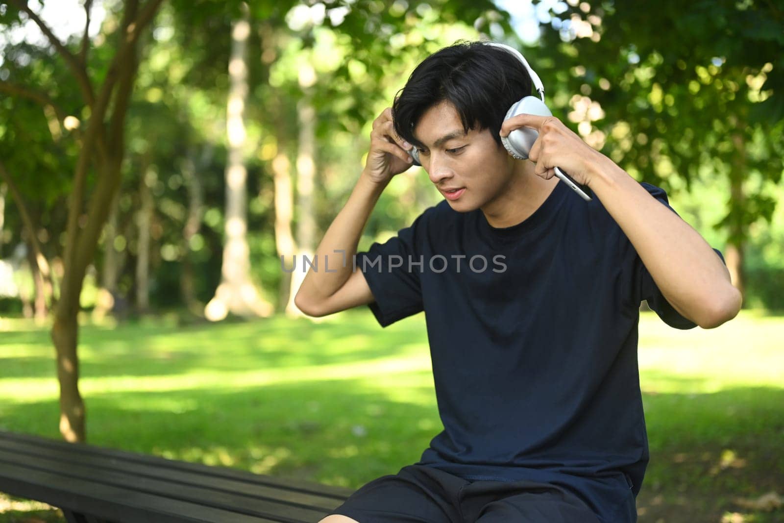 Handsome male runner listening to music on headphones and taking a break after jogging in the park.