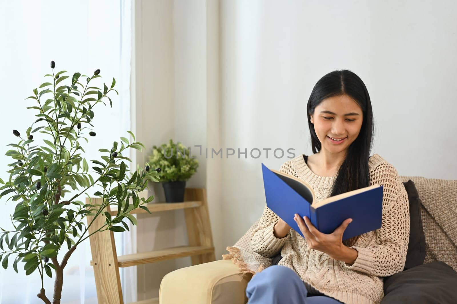 Smiling young woman reading book on sofa at home.