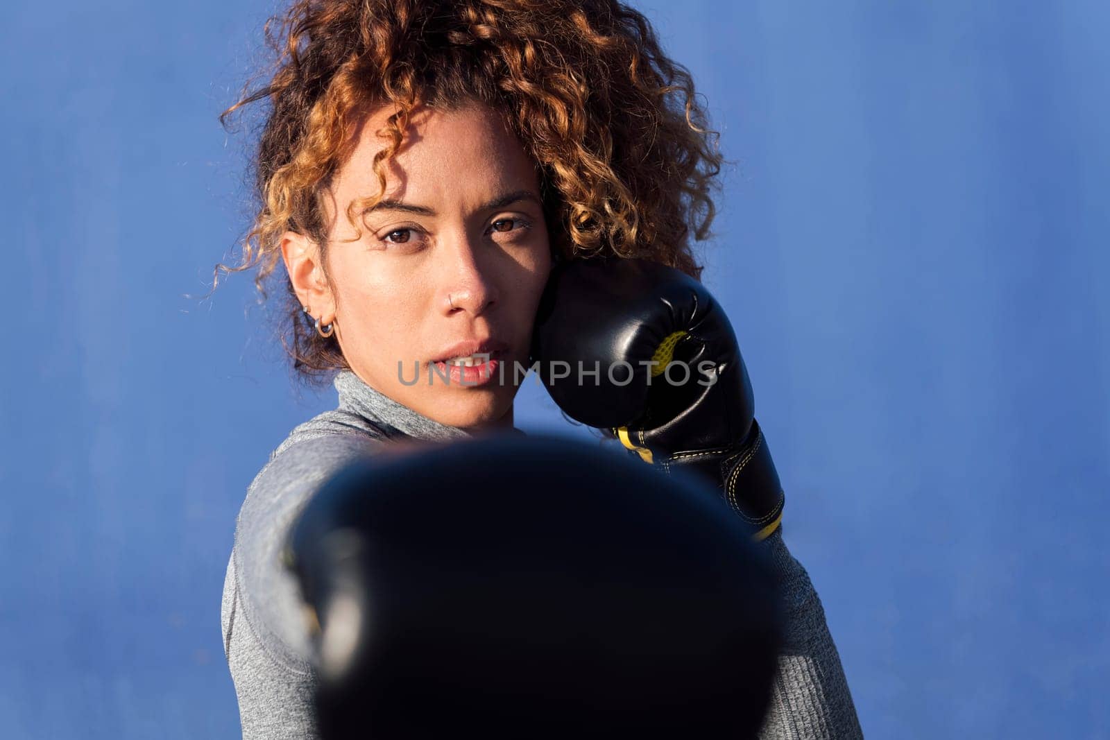 determined adult female boxer with curly hair ready to fight on boxing gloves during workout on blue background, concept of strength and healthy lifestyle