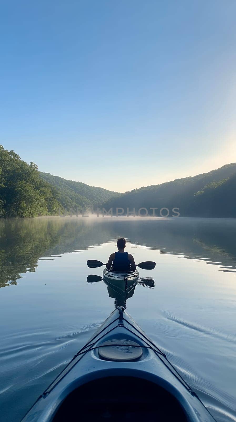 A solo kayaker paddles peacefully on a tranquil lake surrounded by misty hills in the early morning light.