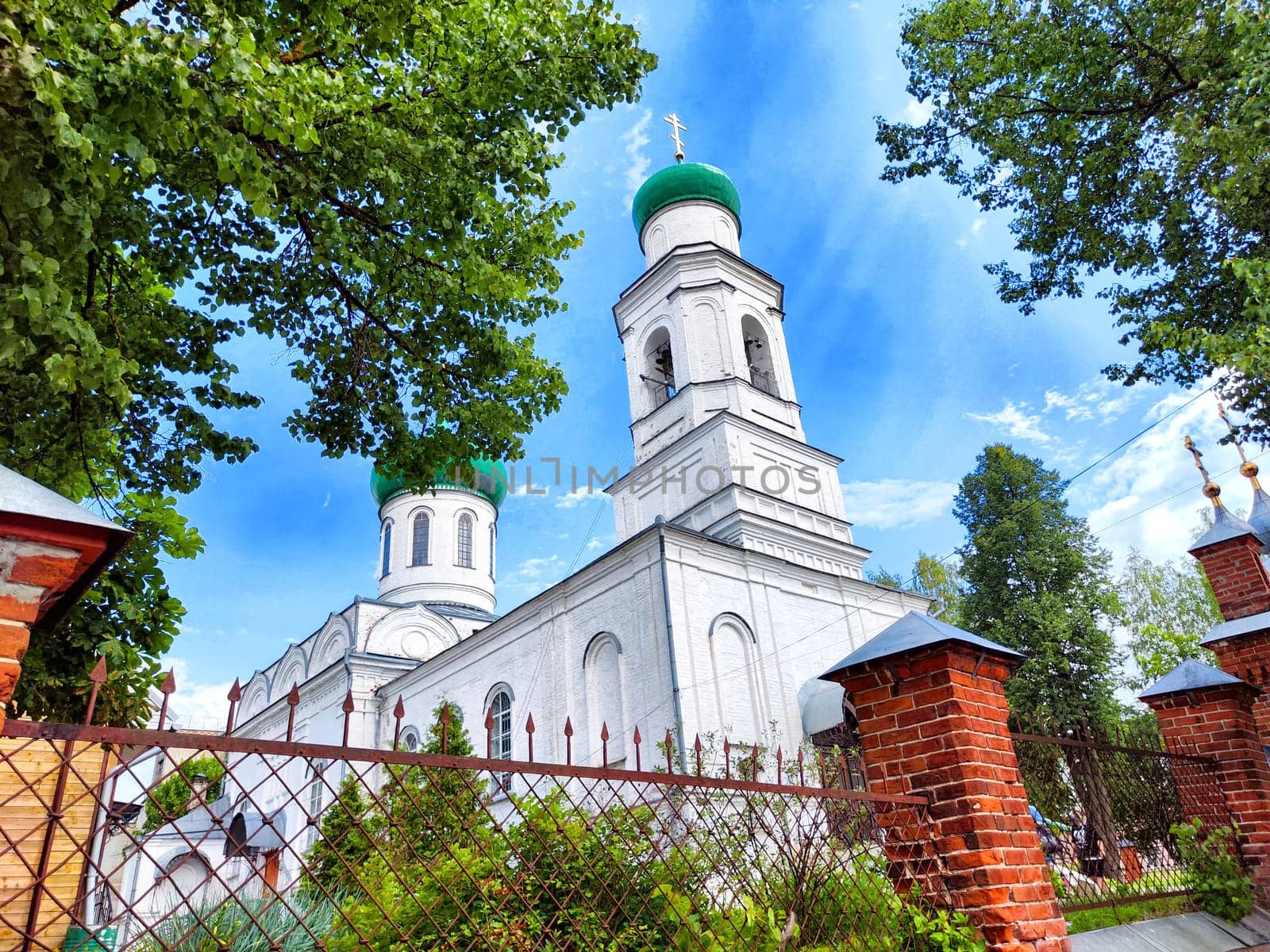 A serene church featuring green domes is nestled among lush trees, contrasting against a bright blue sky.