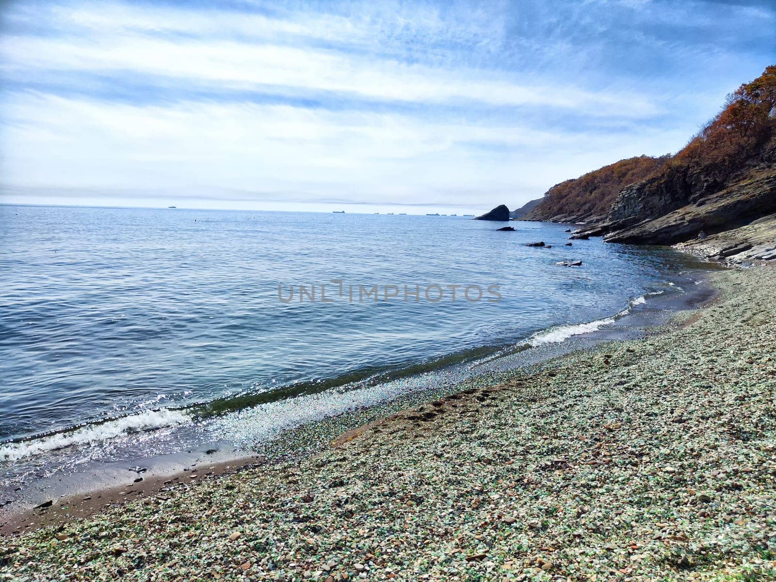 Waves gently lap against a pebbly beach under a clear blue sky near rocky cliffs.