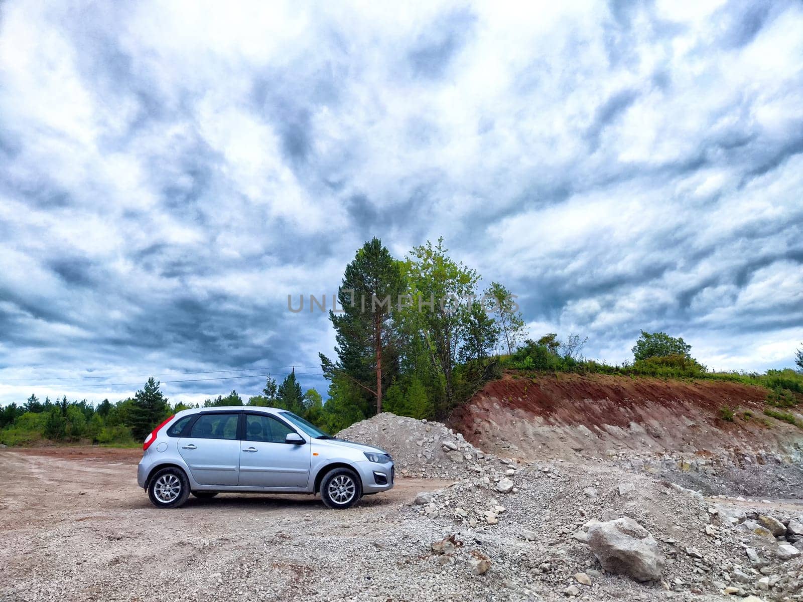 A silver vehicle stands on rocky ground surrounded by greenery and a stunning cloud-filled sky on a sunny day.