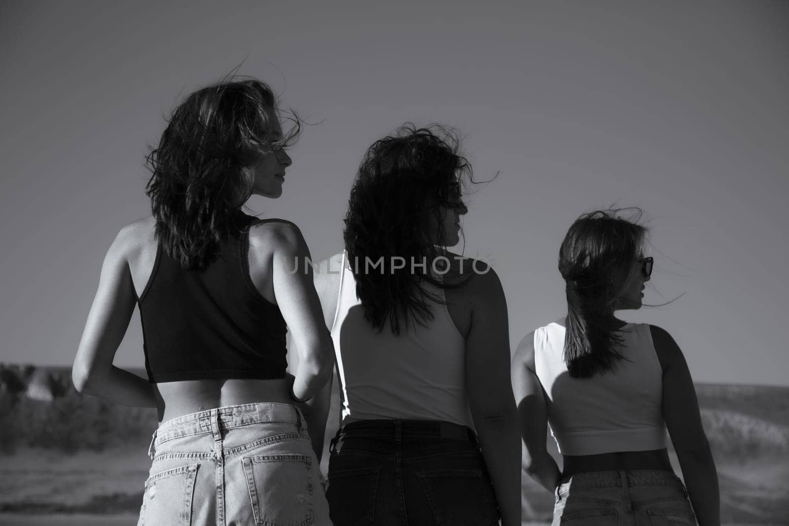 Black and white photo. Three young attractive women stand with their backs turned and their heads turned in profile.
