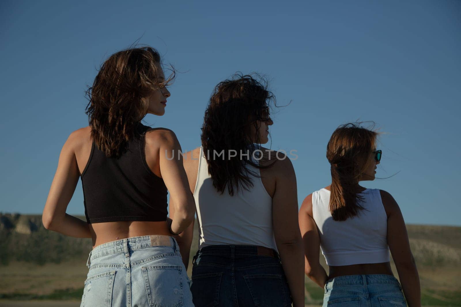 Three young attractive women stand with their backs turned and their heads turned in profile.