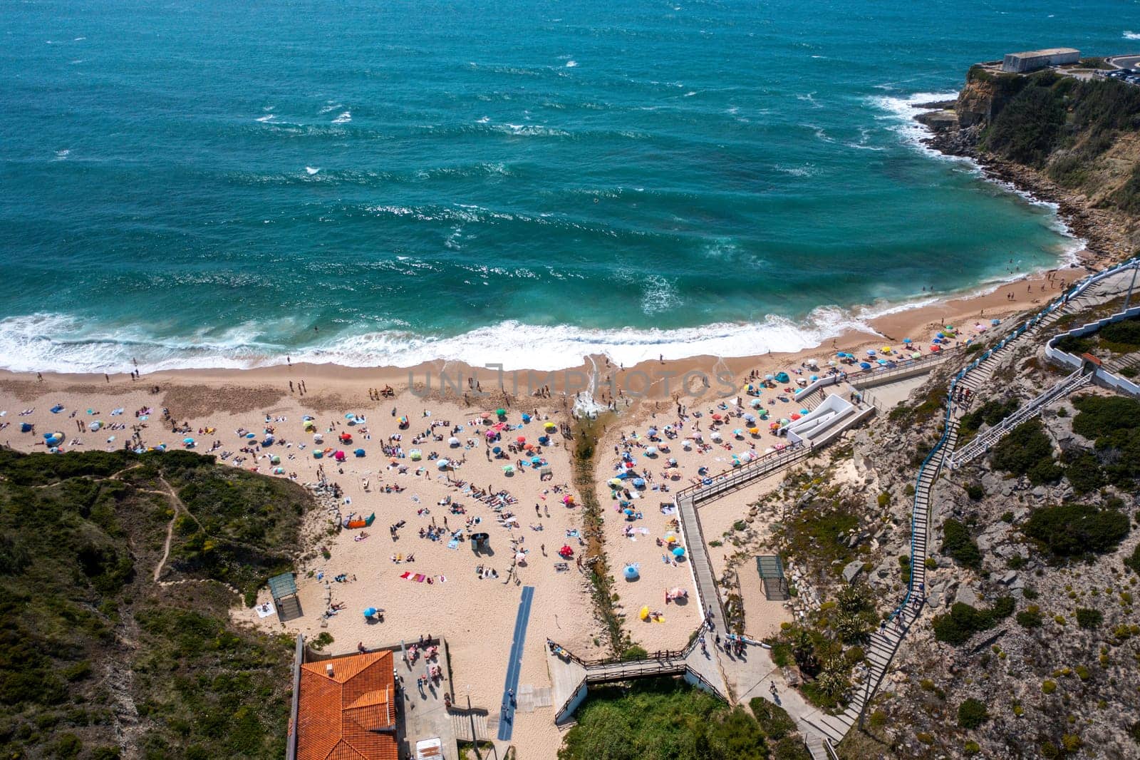 Overhead drone view of anonymous people and parasols on sandy seashore at Magoito Beach. From above of famous destination in Sintra, Portugal during summer.