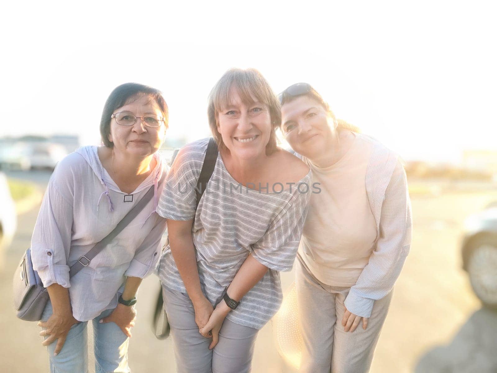 Three friends stand close together with bright smiles, enjoying sunny evening outdoors in a lively parking area