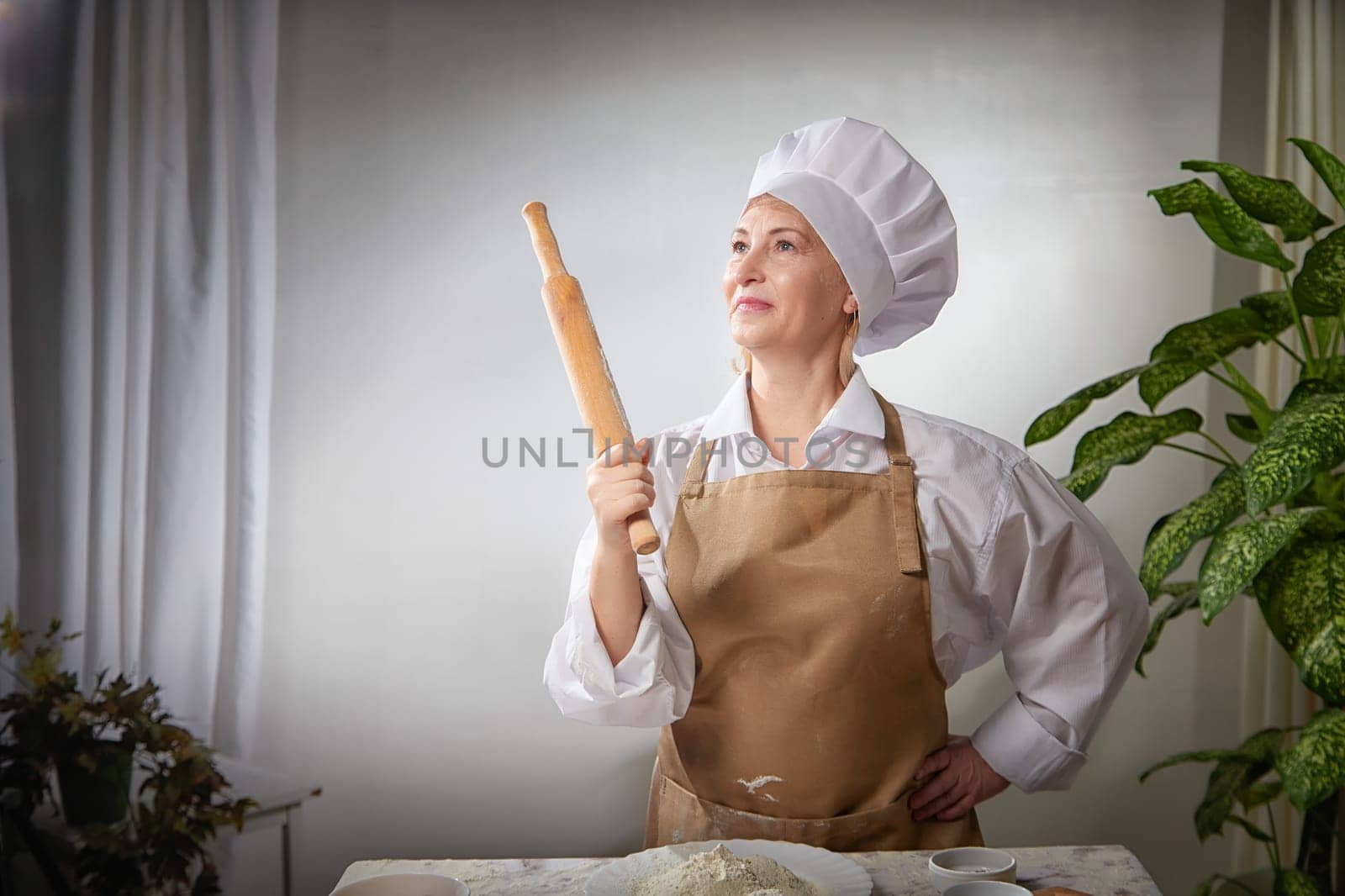 An elderly woman humorously holds a rolling pin, showcasing her passion for cooking with flour in a bright kitchen.